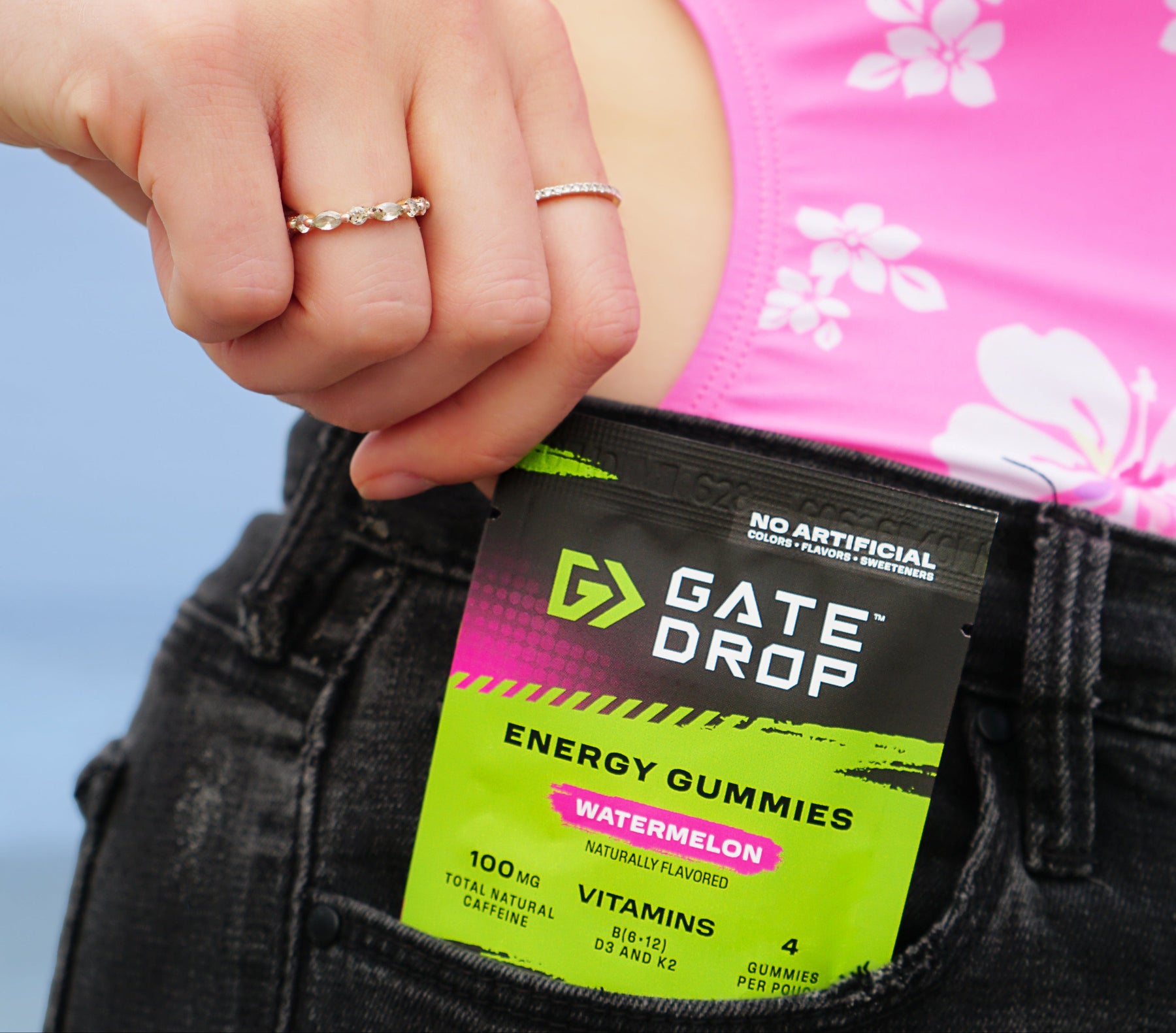 A close-up lifestyle shot of a person wearing a pink floral top and black jeans, pulling a Gate Drop Energy Gummies packet out of their front pocket. The packet is watermelon-flavored and features a black, pink, and lime green design.
